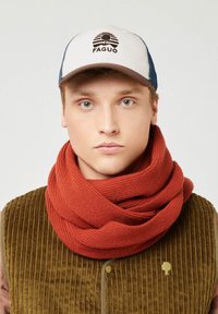 Jonge man met een wit en blauwe pet met een boomlogo, een dikke roestkleurige gebreide sjaal en een bruine ribfluwelen vest met een boomembleem.