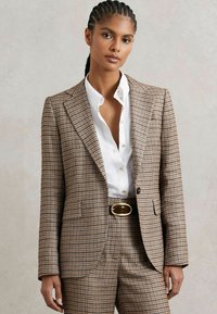 Hnedý blazer s kockovaným vzorom a štruktúrovaným dizajnom, klopami a dvoma prednými vreckami, skombinovaný s bielou košeľou s gombíkmi a opaskom.