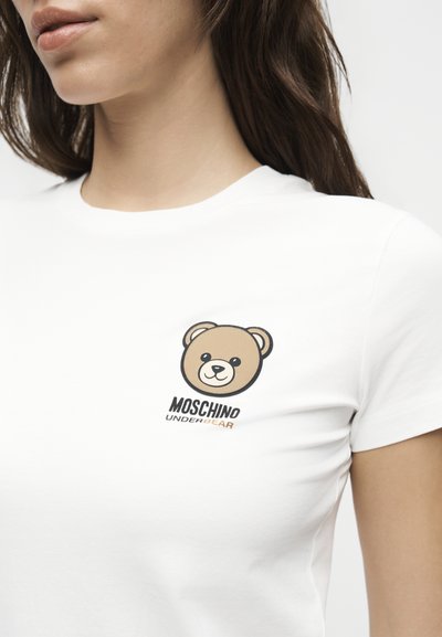 Γυναίκα που φοράει λευκό T-shirt με μικρό καρτούν αρκουδάκι στο στήθος και το κείμενο "MOSCHINO UNDERBEAR".