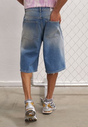 Mann, der ausgewaschene blaue Jeansshorts und weiße Sneaker trägt, steht mit einer Hand in der Gesäßtasche an einem Maschendrahtzaun.