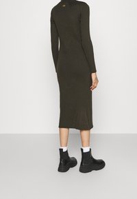 Kvinna som bär en ribbad, långärmad olivgrön midiklänning med svarta ankelboots och vita strumpor, stående vänd bortåt mot en enfärgad bakgrund.