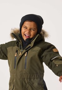 Green winter jacket with a fur-lined hood, featuring two zippered pockets and tan leather accents. Worn with a black beanie and scarf.