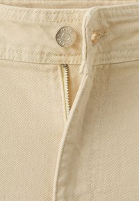 Jeans en denim beige avec fermeture éclair partiellement ouverte et bouton en métal gravé "MNG JEANS" sur la ceinture.