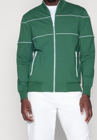 Giacca verde con zip realizzata in materiale leggero, con strisce bianche attraverso il petto e due tasche frontali con zip. Stile con colletto alto.