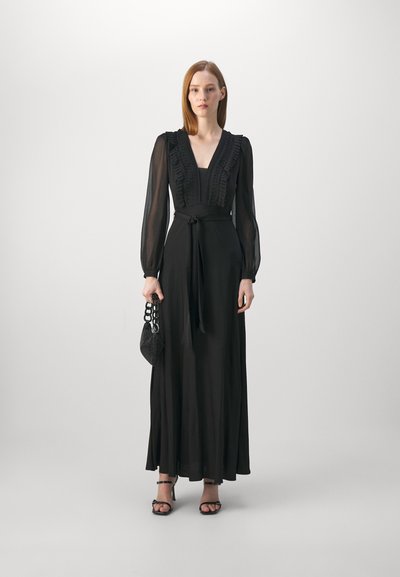 Robe noire longue avec des manches longues transparentes, un col froncé et une taille cintrée. Le mannequin tient un sac noir texturé avec une anse en chaîne.