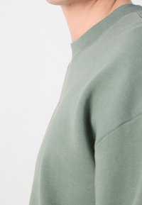 Primo piano di una persona che indossa un maglione a girocollo verde chiaro, che mostra collo, spalla e parte superiore del petto su uno sfondo semplice.