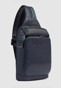 Bolso bandolera de cuero azul marino con textura suave, múltiples bolsillos con cremallera y una correa de hombro acolchada. Destaca el logotipo de Piquadro de manera prominente.