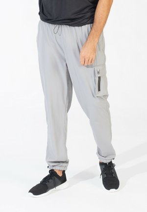 Pantalons cargo gris clair en matériau synthétique avec une taille élastique, une poche zippée et des poignets resserrés. Associés à des baskets noires.