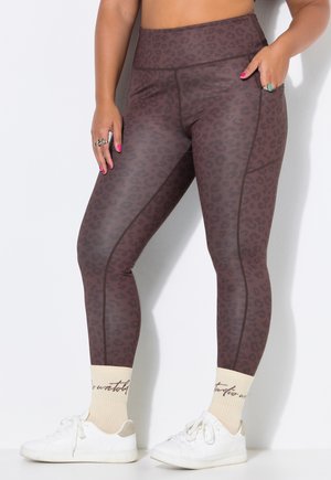 Vrouw draagt hoge taille bruine legging met luipaardprint en zijzak, witte sneakers en crèmekleurige sokken met cursieve tekst.