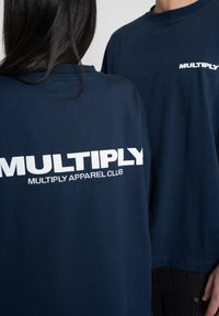 T-pait sinimusta puuvillaa, joissa on suuret valkoiset painetut tekstit: "MULTIPLY" ja "MULTIPLY APPAREL CLUB" selässä. Lyhyet hihat ja rento malli.