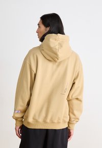 Felpa beige con cappuccio regolabile, caratterizzata da disegni in linea e testo colorato sulla manica. Tessuto morbido con vestibilità rilassata.