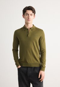 Olivno zelena dolga polo majica s kragnico, tremi gumbi in vezenim logotipom. Gladka tekstura in prilegajoč dizajn. Nosi se z temno modrimi jeans hlačami.