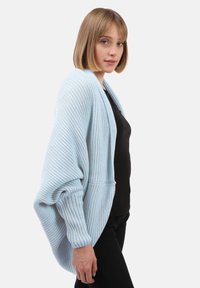 Giovane donna con capelli biondi corti che indossa un cardigan voluminoso di colore azzurro chiaro sopra una maglietta nera e pantaloni neri, in piedi contro uno sfondo bianco.