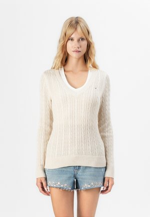 Donna bionda che indossa un maglione a V in crema con lavorazione a trecce sopra una maglietta bianca e pantaloni shorts in denim ricamati, in piedi contro uno sfondo bianco uniforme.
