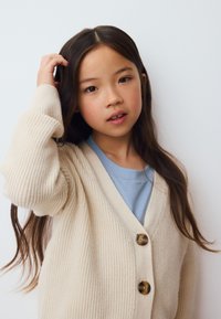 Cardigan de canalé color crema con escote en V, con dos grandes botones de carey. Llevado sobre una camisa azul claro, con cabello largo y oscuro visible.