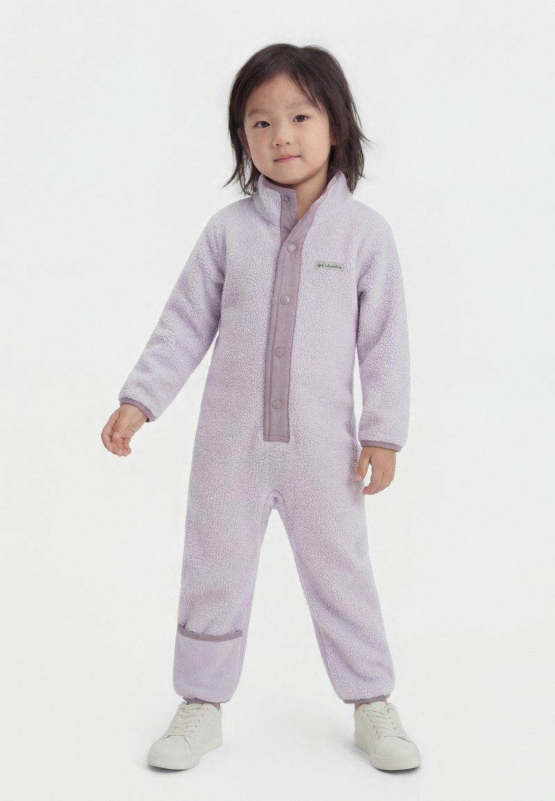 Gyermek áll, világoslila, bolyhos egyrészes kezeslábasban, elöl patentos és fehér tornacipőben, egy sima világos háttér előtt.