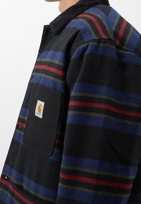 Stripet flannel skjorte i sort, blå, grøn og rød, med en sort påsyet lomme og et fremtrædende logo. Har krave og knapdetaljer.