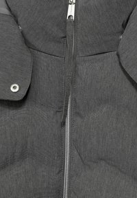 Grå vadderad jacka med en slät, texturerad yta. Har en framdragkedja, tryckknappsstängning på kragen och en etikett med varumärkesinformation.