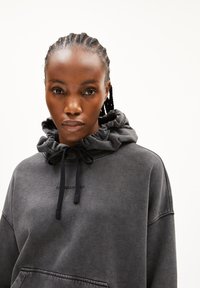 Eine Frau mit geflochtenem Haar, die einen dunkelgrauen Hoodie mit Kordeln trägt, schaut direkt in die Kamera vor einem schlichten, hellen Hintergrund.