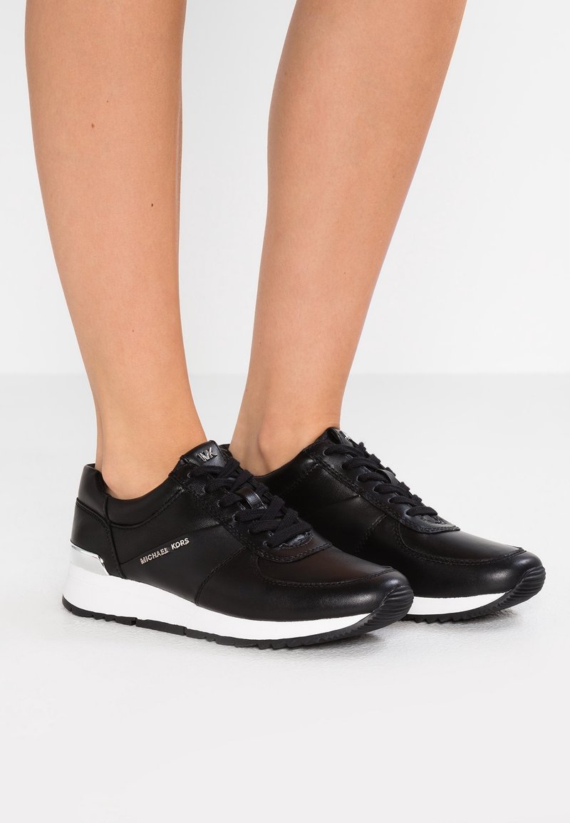 Baskets en cuir noir Michael Kors avec semelles blanches portées pieds nus, vues de profil sur un fond uni.