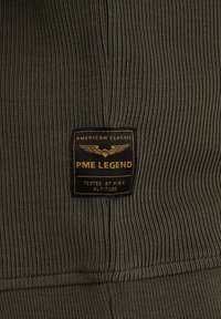 Tela acanalada de color verde oliva oscuro con una etiqueta negra y dorada. El texto dice "PME LEGEND" y "TESTED AT HIGH ALTITUDE."