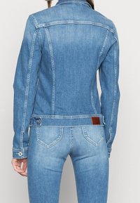 Vaaleansininen farkkutakki, jossa on istuva muotoilu, klassinen kaulus ja nappikoristeet. Takissa on ruskea nahkapatchi vyötäröllä.