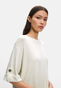Sweter w kolorze kremowym, dziany, z krótkim rękawem, z guzikowym detalem przy mankiecie. Miękka tekstura i luźny krój.