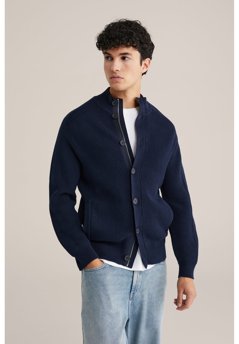 Marinblå stickad kofta med hög krage, knäppning och ribbad struktur. Inkluderar två framfickor och kombineras med en vit skjorta.