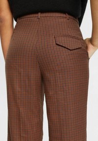 Pantalon petit à carreaux marron et noir avec une poche à rabat à l’arrière, porté avec un haut noir, montré de la taille vers le bas.