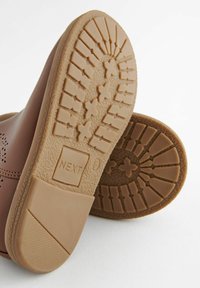 Chaussures flexibles brunes avec une semelle en caoutchouc texturée. Présente un motif de semelle et une petite étiquette "NEXT". Le matériau supérieur est perforé.