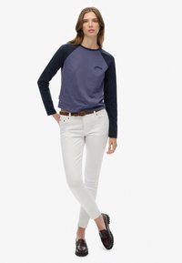 Vrouw in een blauwe langekleurige raglan shirt, witte slim-fit broek, een bruine gevlochten riem en donkerbruine loafers, staand met gekruiste benen.