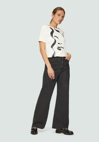 T-shirt blanc avec un design de visage abstrait noir, associé à un pantalon large taille haute noir et des chaussures plates noires. Texture en coton lisse.