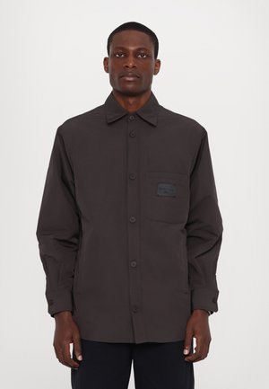Camisa de botão cinza escuro com um bolso no peito contendo uma etiqueta, mangas longas e um corte descontraído. Tecido liso e leve.