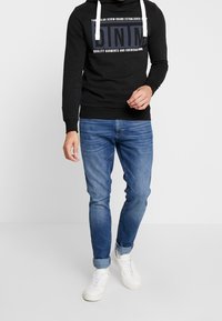 Czarna bluza z kapturem z białymi sznureczkami i dużym grafiką z przodu. W zestawie z jasnoniebieskimi dżinsami i białymi sneakersami. Luźny, dopasowany krój.