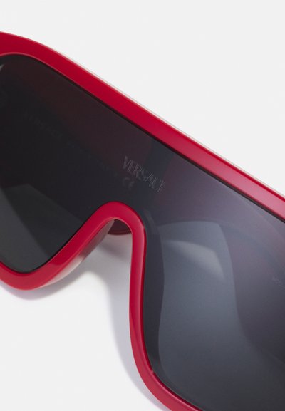 Ochelari de soare roșii, supradimensionați, cu lentile întunecate, design curbat și logo-ul Versace gravat lângă balama. Fabricati din plastic lucios.