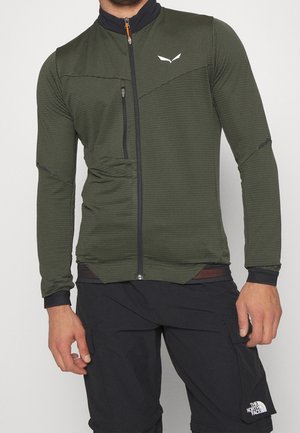 Mężczyzna ubrany w oliwkowo-zieloną kurtkę na zamek błyskawiczny z kieszenią na piersi oraz czarne spodnie z logo The North Face na prawej udzie.