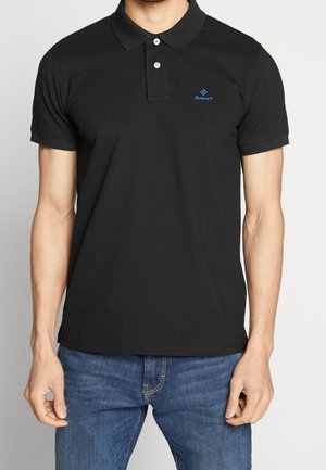 Homme portant un polo noir à manches courtes avec deux boutons blancs et un petit logo GANT bleu, associé à un jean bleu.