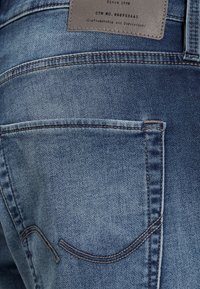 Modré denimové džíny s vybledlým povrchem, které mají zadní kapsu, kontrastní švy a koženou etiketu na pase s textovými údaji.