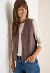 Lächelnde Frau in einem weichen, braunen ärmellosen Weste über einem langärmeligen cremefarbenen Hemd und blauen Jeans, die vor einer weißen Wand steht.