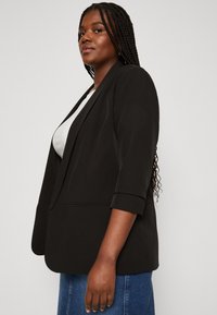 Black blazer with a structured design, three-quarter sleeves, and two front pockets. Worn over a white top, paired with blue denim.