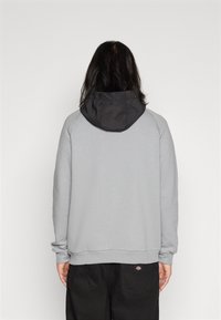 Felpa grigia con cappuccio scuro, maniche raglan e polsini a coste. Presenta un logo in basso a destra. Texture liscia, design casual.