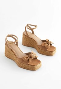 Braune Wildleder-Wedge-Sandalen mit geflochtenem Jute-Mittelsohle, quadratischer Zehenpartie und dekorativem KnotenDetail am vorderen Riemen. Verstellbarer Knöchelriemen inklusive.