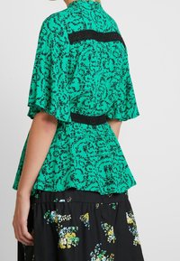 Top peplum verde con patrón floral negro, mangas cortas y una cintura texturizada negra. Silueta en línea A con detalle de tela en capas.