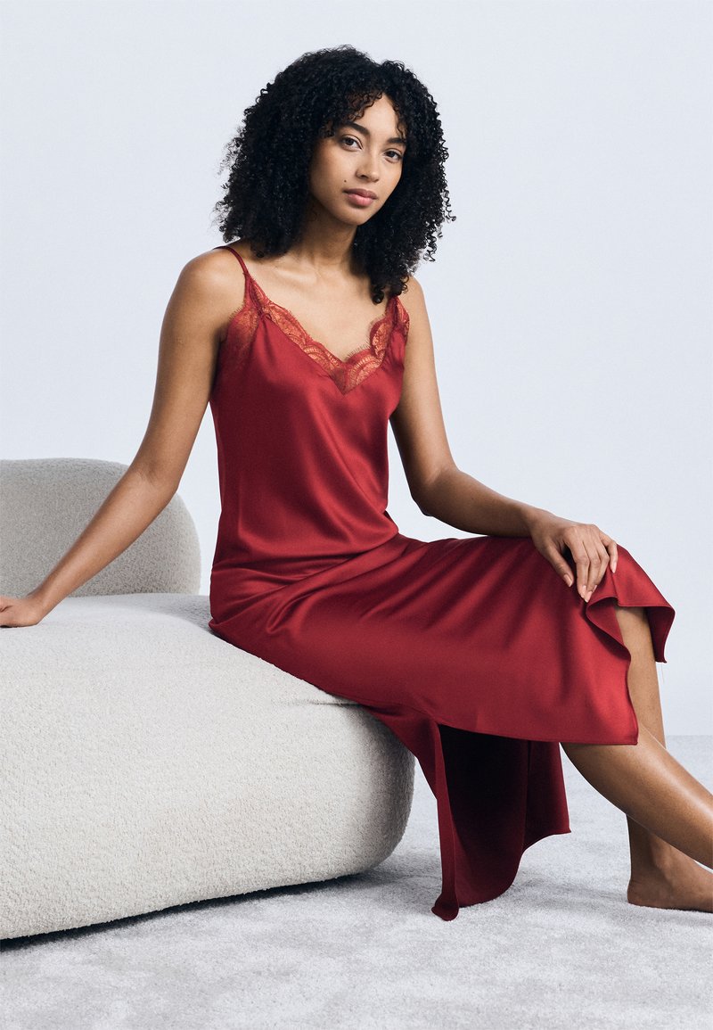 Γυναίκα με σγουρά μαλλιά που κάθεται σε ένα λευκό πάγκο με υφή, φορώντας ένα κόκκινο σατέν φόρεμα ολίσθησης με δαντέλα, μπροστά σε ένα απλό ανοιχτόχρωμο φόντο.