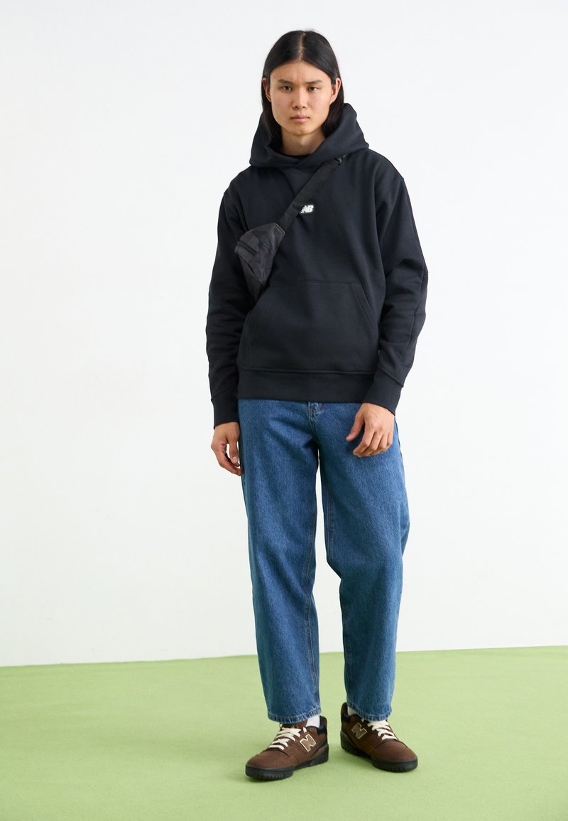 Sudadera con capucha negra y bolsillo frontal, combinada con unos vaqueros azules holgados y zapatillas marrones. Se lleva un pequeño bolso cruzado negro.