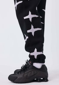 Los pantalones de chándal negros presentan patrones en forma de estrella blanca y puños fruncidos. Se llevan unas zapatillas Nike negras con detalles blancos y paneles texturizados.