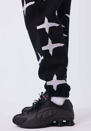 Pantaloni della tuta neri con motivi bianchi a forma di stella e polsini arricciati. Scarpe Nike nere con accenti bianchi e pannelli texture.