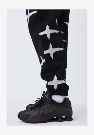 Zwarte trainingsbroek met witte stervormige patronen en vergaarde boorden. Zwarte Nike schoenen met witte accenten en textured panelen worden gedragen.