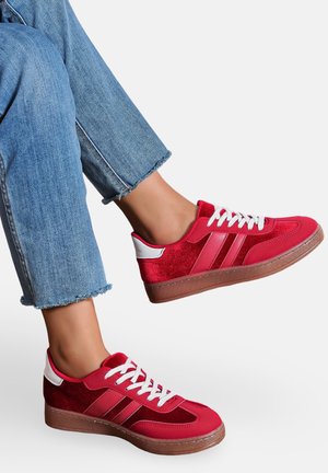 Jean bleu longueur cheville portés avec des baskets rouges à lacets blancs et semelles marron, sur fond blanc.