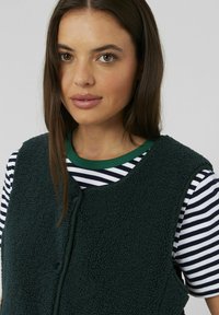 Jeune femme aux cheveux bruns raides, portant un gilet en polaire noir par-dessus une chemise à rayures blanches et noires à manches courtes, sur un fond uni.